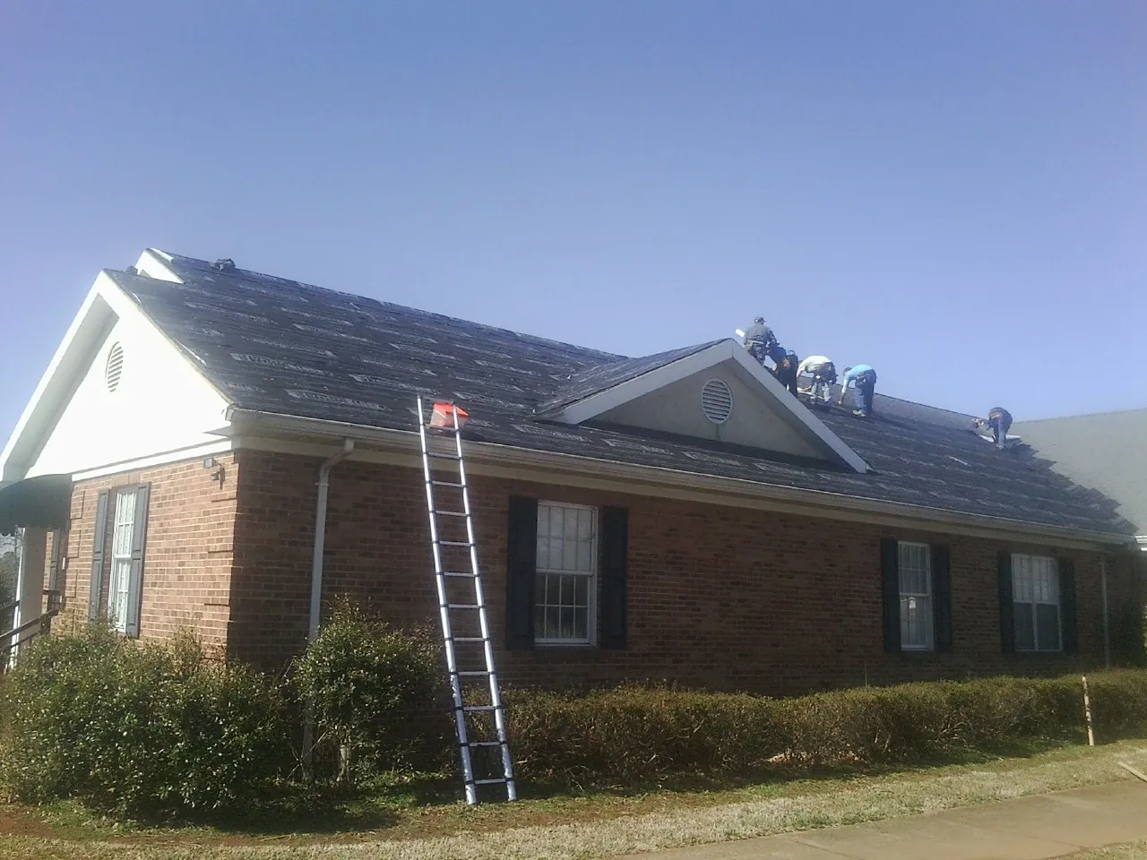 Crew tearing shingles off a brick ranch roof with a ladder set — Emergency Roof Repair work by our crew in Northborough