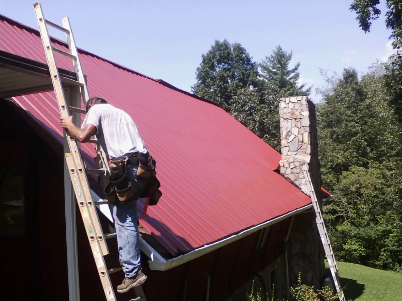 Roofer on a ladder beside a red metal roof and stone chimney — Soffit & Fascia Repair by Roofing Brothers in Northborough